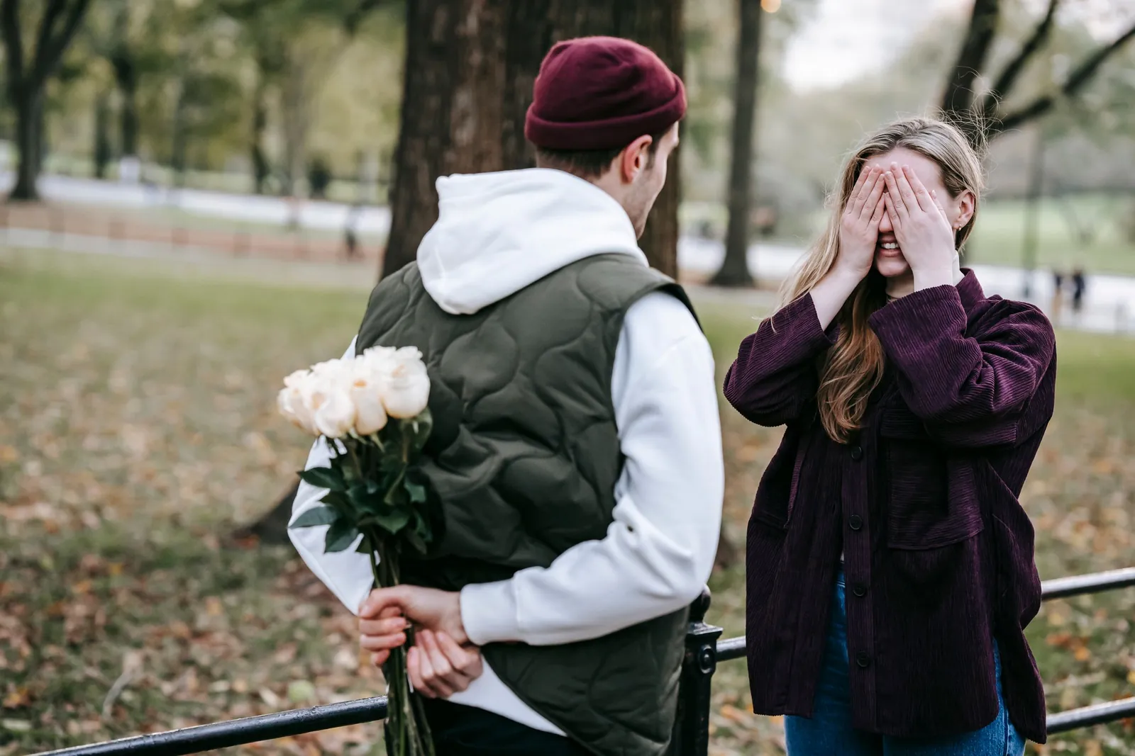 He hides a bouquet of white roses while she covers her eyes, ready for the surprise