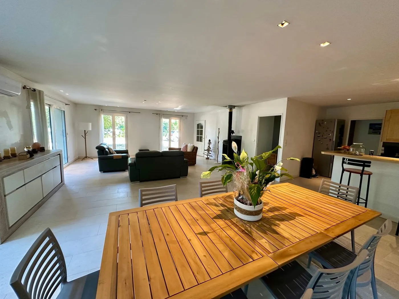 La salle de 75 m² du Domaine du Dahu : grande table en bois, poêle à bois, cuisine ouverte et baies vitrées